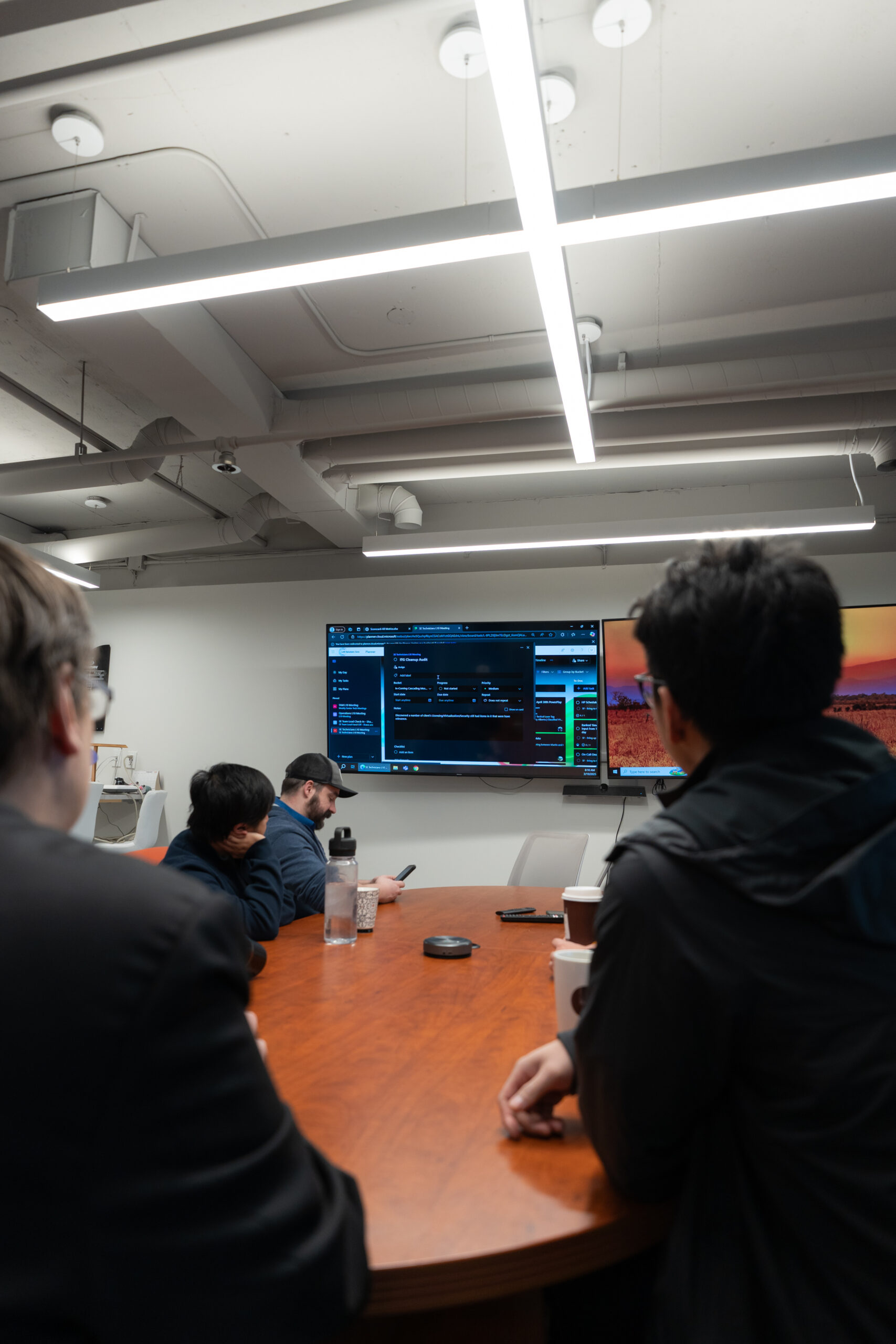 A group of professionals seated around a boardroom table watching a large screen displaying data charts and computer code, representing teamwork and cybersecurity monitoring.